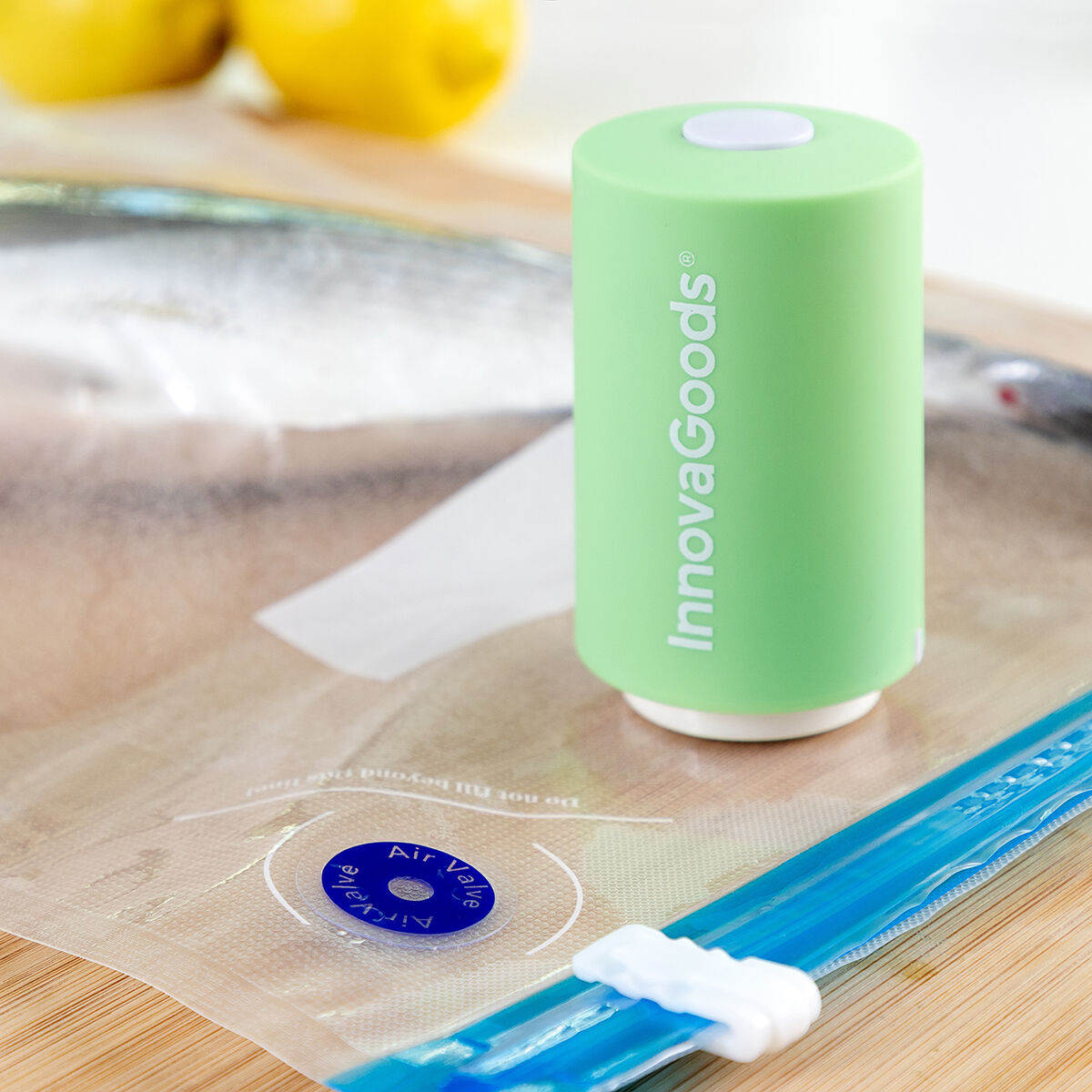 Una sigillatrice sottovuoto InnovaGoods verde con la parte superiore bianca. Un sacchetto sigillato, cerniera blu e due limoni sullo sfondo, su legno.