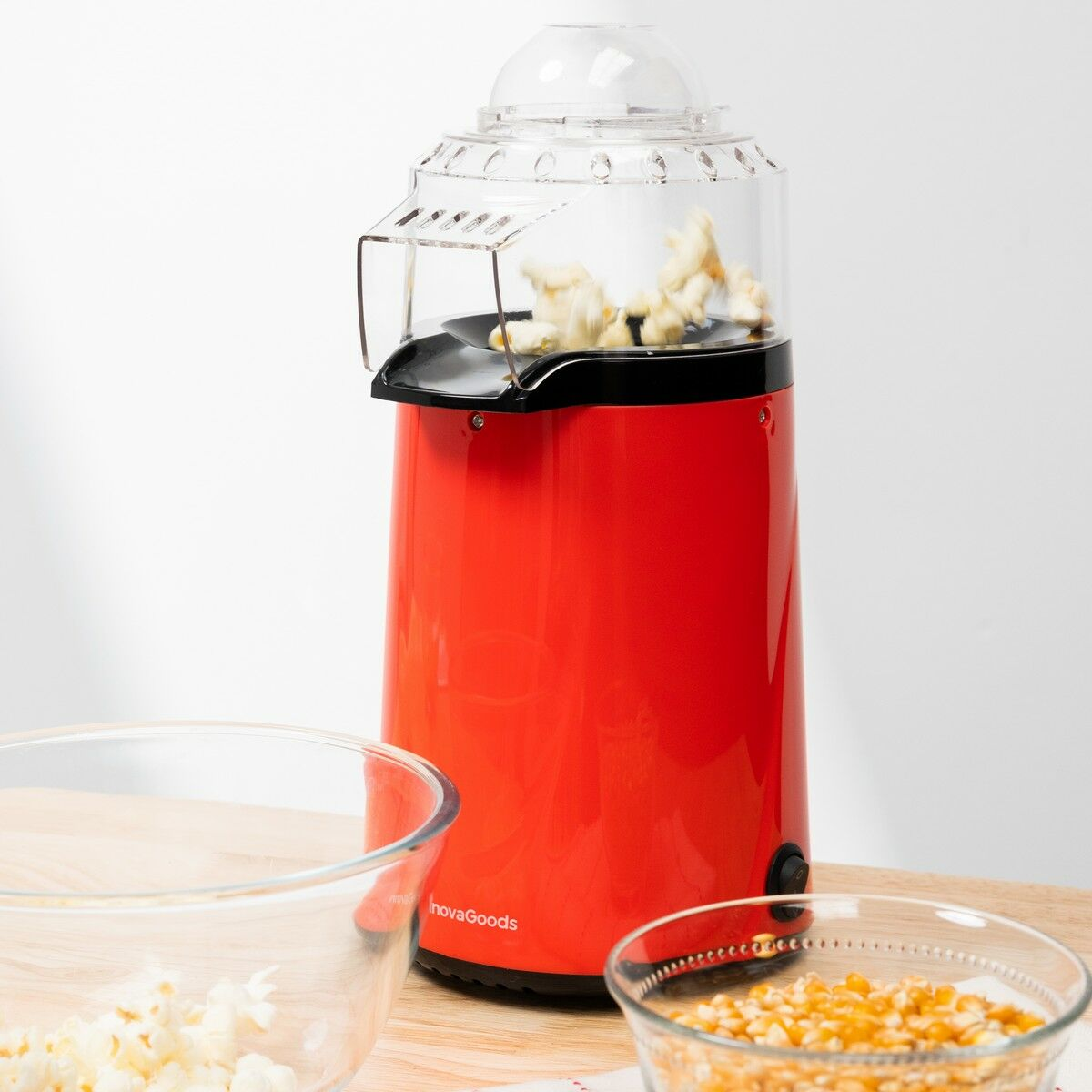 Ein roter Popcornbereiter mit Popcorn im Inneren, zwei Glasschalen mit Popcorn und Körnern auf einem Holztisch. Weißer Hintergrund.