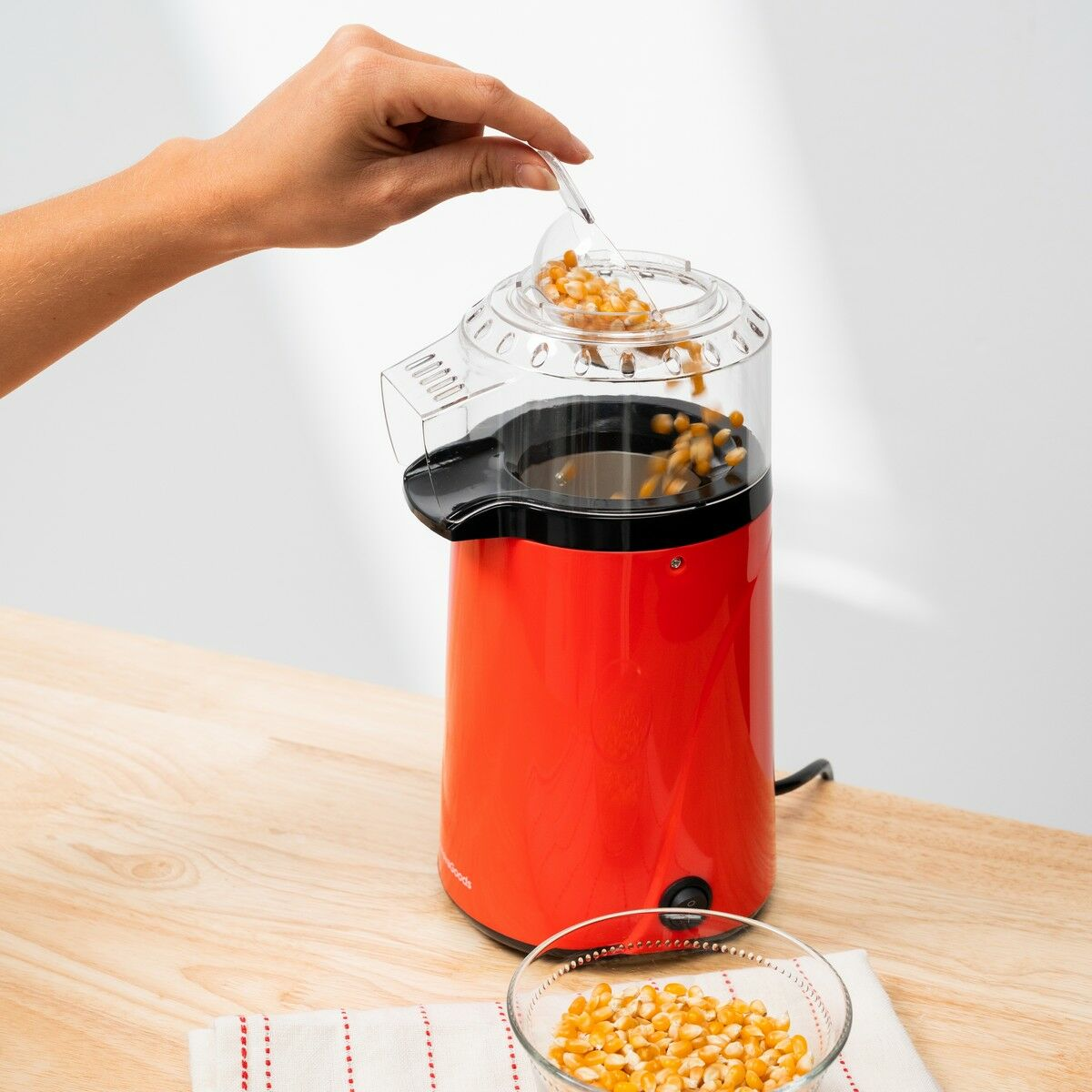 Eine Hand schüttet Maiskörner in eine rote Popcornmaschine. Eine Schüssel mit Körnern steht auf einem Tisch. Der Hintergrund ist weiß.