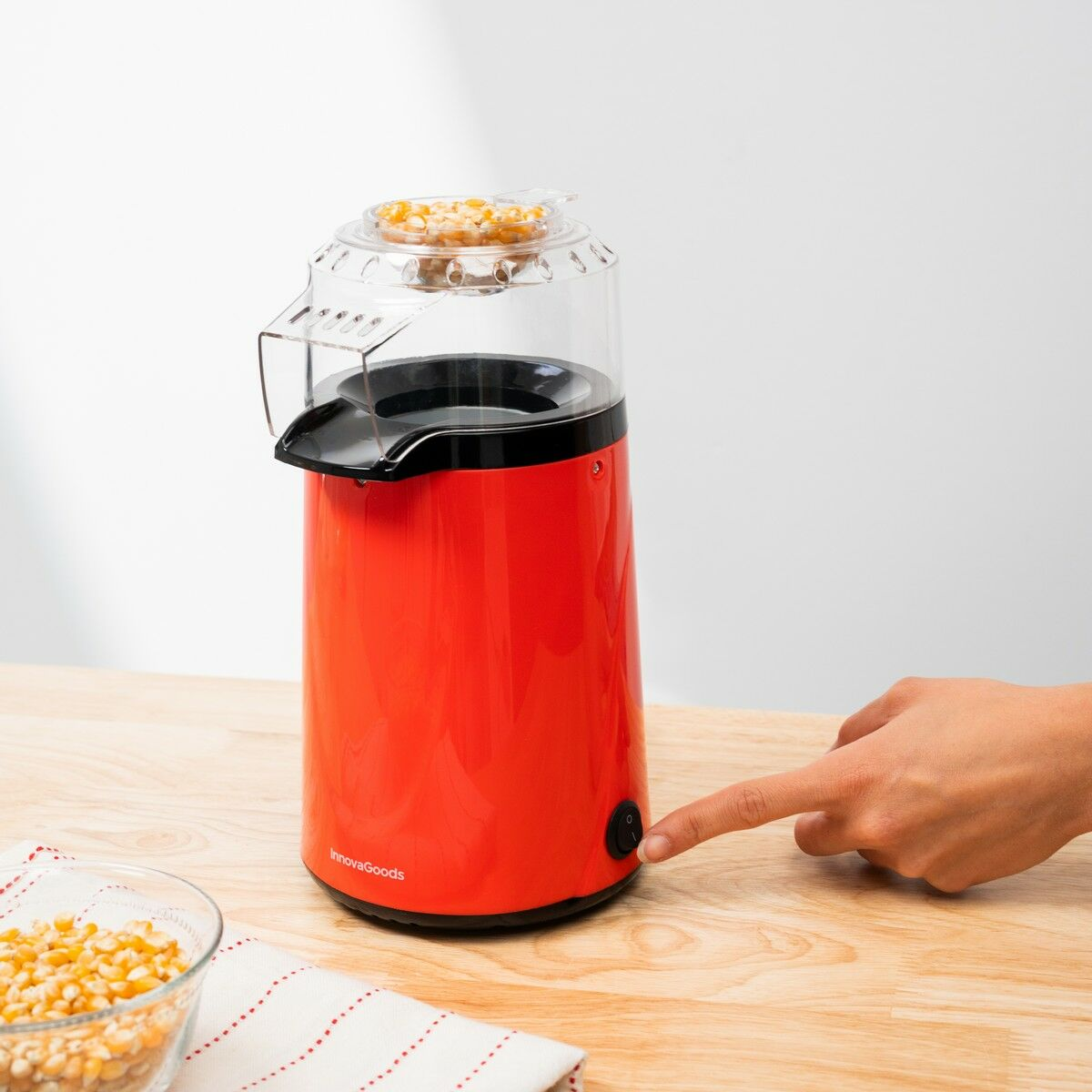Ein roter InnovaGoods Popcornautomat steht auf einem Holztisch. Eine Hand drückt den Einschaltknopf. Mais und eine Schüssel sind im Vordergrund.