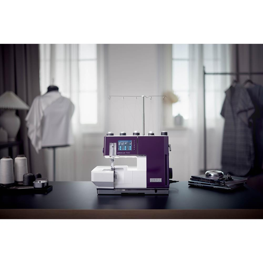 Una máquina de coser morada y blanca sobre una mesa. Un maniquí con una blusa blanca a la izquierda. Cortinas y una ventana al fondo.