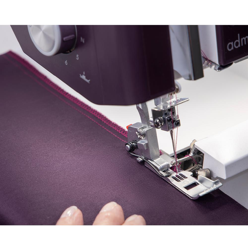 Una máquina de coser cose tela morada. La máquina es de color púrpura oscuro y plateado. La tela tiene costuras rosas en el borde.