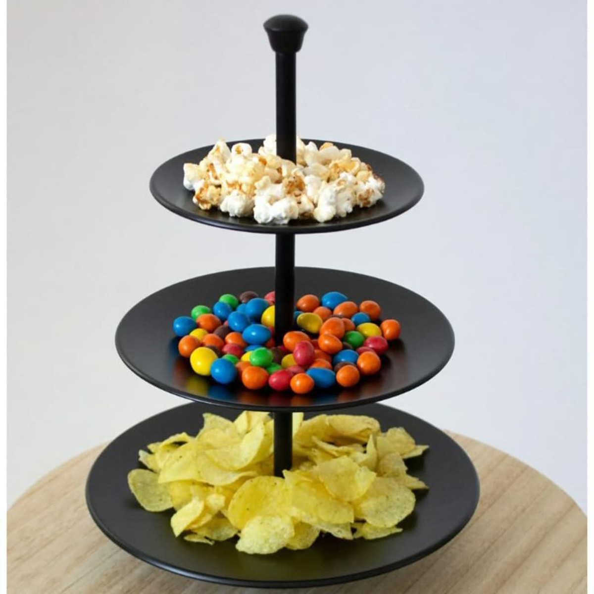 Ein dreistufiges, schwarzes Serviertablett mit Popcorn, bunten Süßigkeiten und Kartoffelchips. Darunter ein Holztisch und ein weißer Hintergrund.