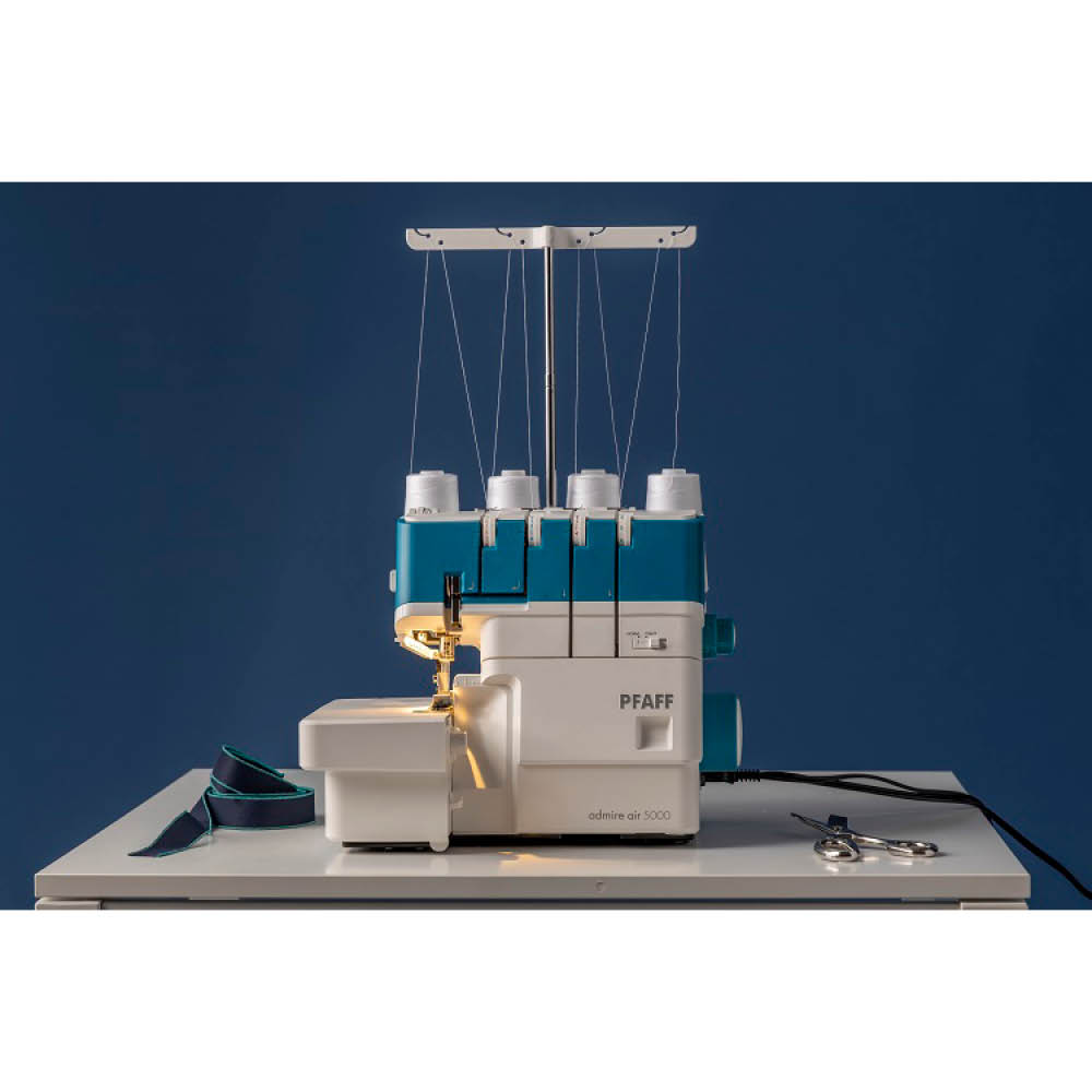 Una máquina overlock Pfaff admire air 5000 blanca y verde azulado está sobre una mesa blanca. Tijeras y tela sobre la mesa, fondo azul.