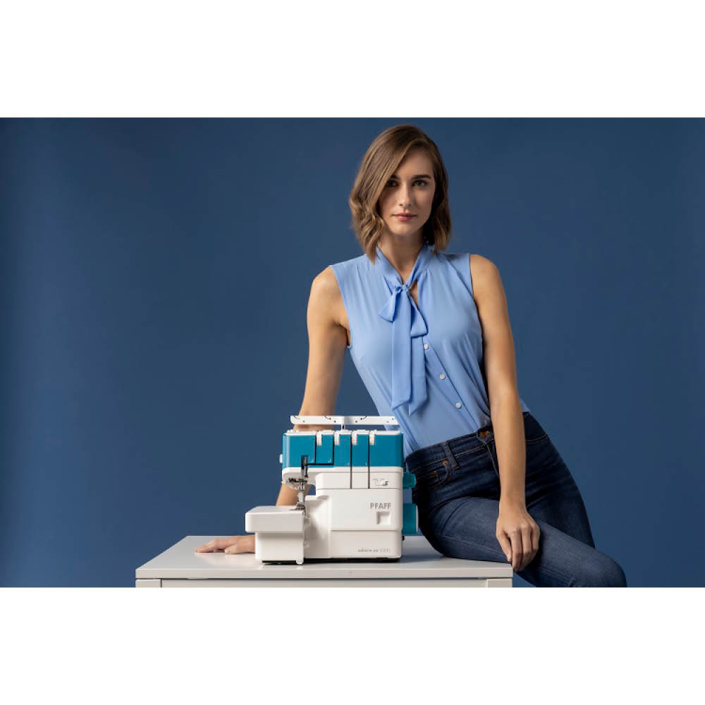 Una mujer con una blusa azul y vaqueros se apoya en una mesa blanca con una máquina de coser. Detrás hay un fondo azul.