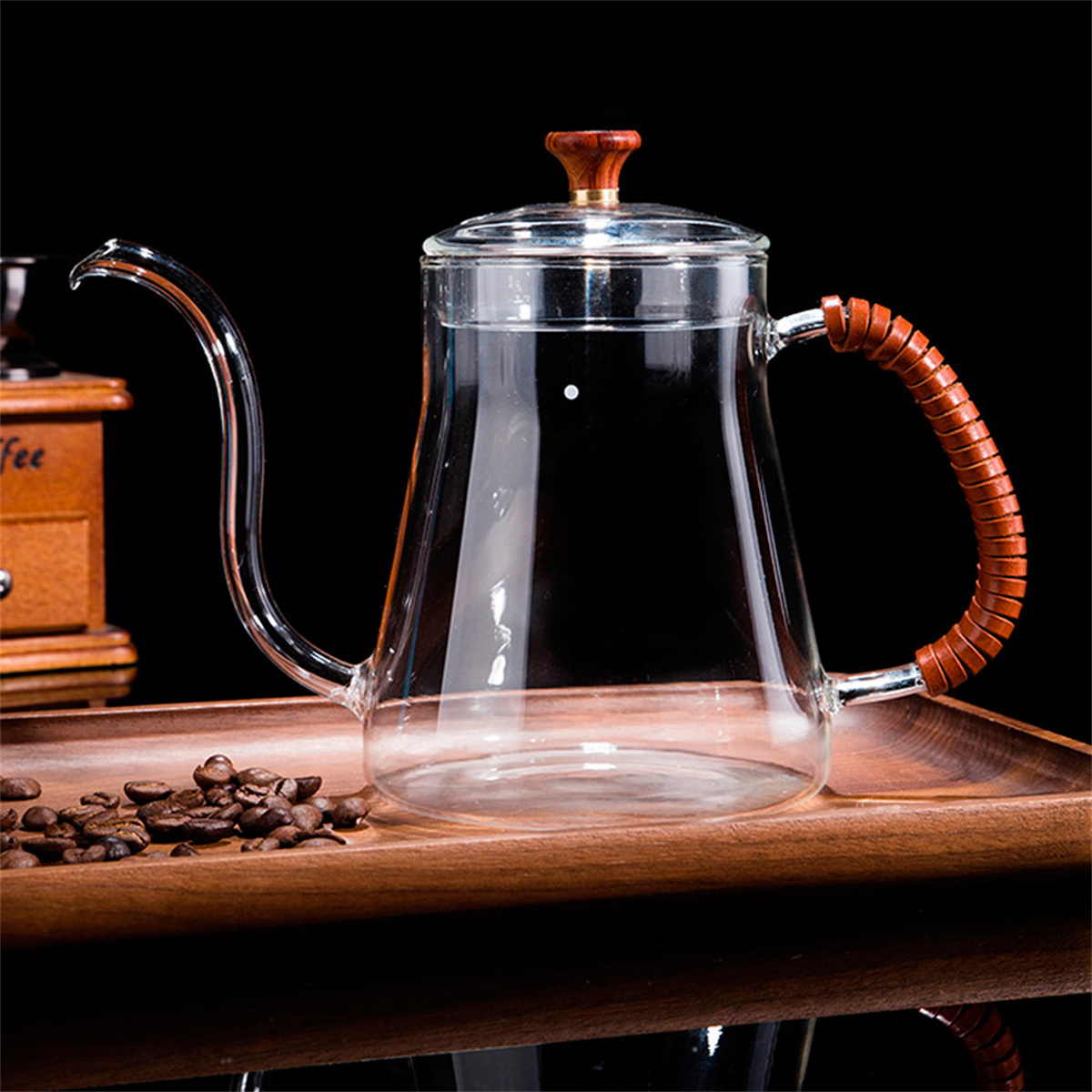 Ein klarer Glaskessel mit Holzgriff und Deckel, gefüllt mit Wasser. Kaffeebohnen und eine Mühle stehen auf einem Holztablett.