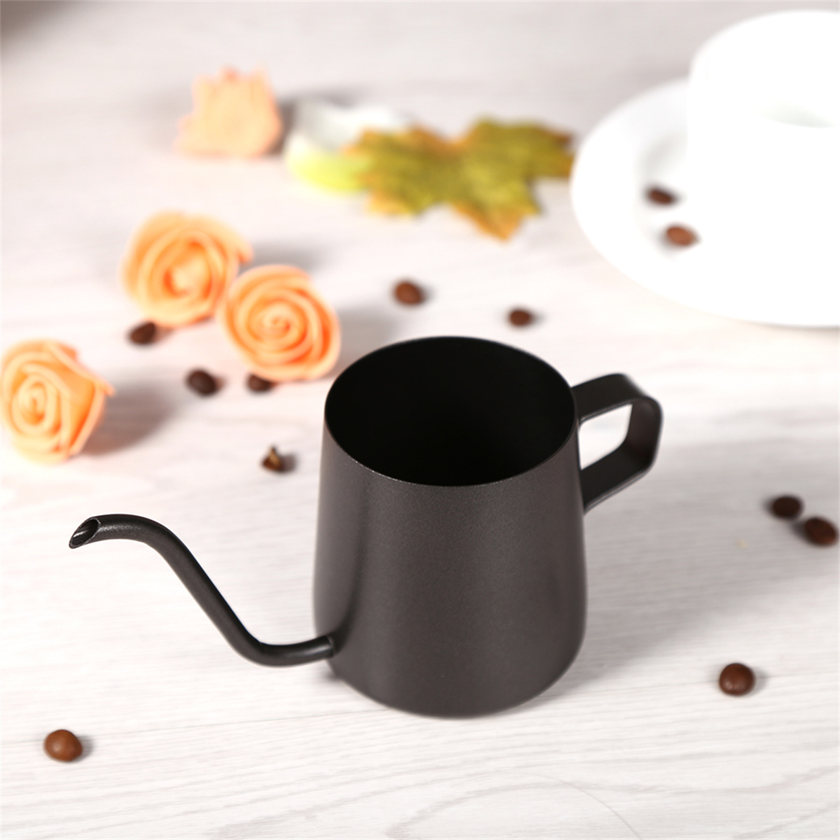 Schwarze Kaffeekanne mit langem Ausguss, umgeben von Kaffeebohnen, orangefarbenen Rosen und einer weißen Tasse auf Holzoberfläche.