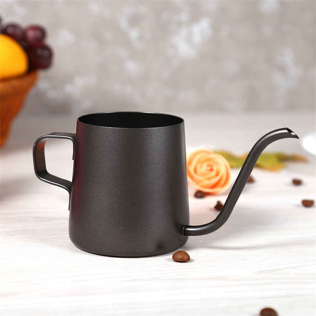 Eine schwarze, metallische Kaffeekanne mit langem Ausguss und Griff. Auf dem Tisch liegen eine Blume und Kaffeebohnen. Im Hintergrund Früchte.