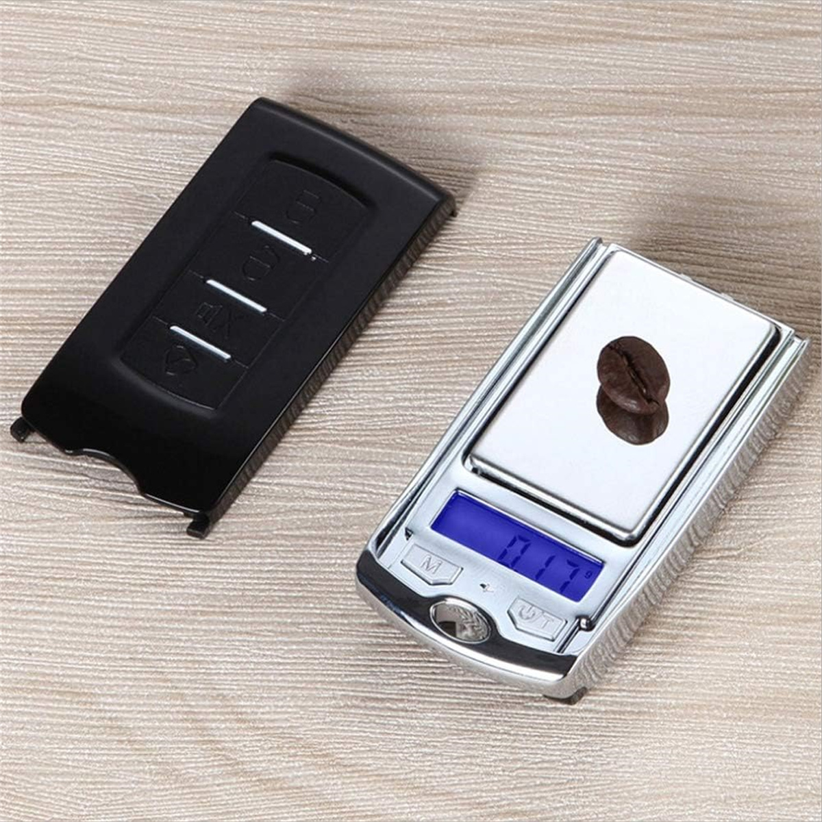 Eine kleine Digitalwaage mit einer Kaffeebohne darauf, neben einem schwarzen Rechteck mit Knöpfen, auf einer Holzoberfläche.