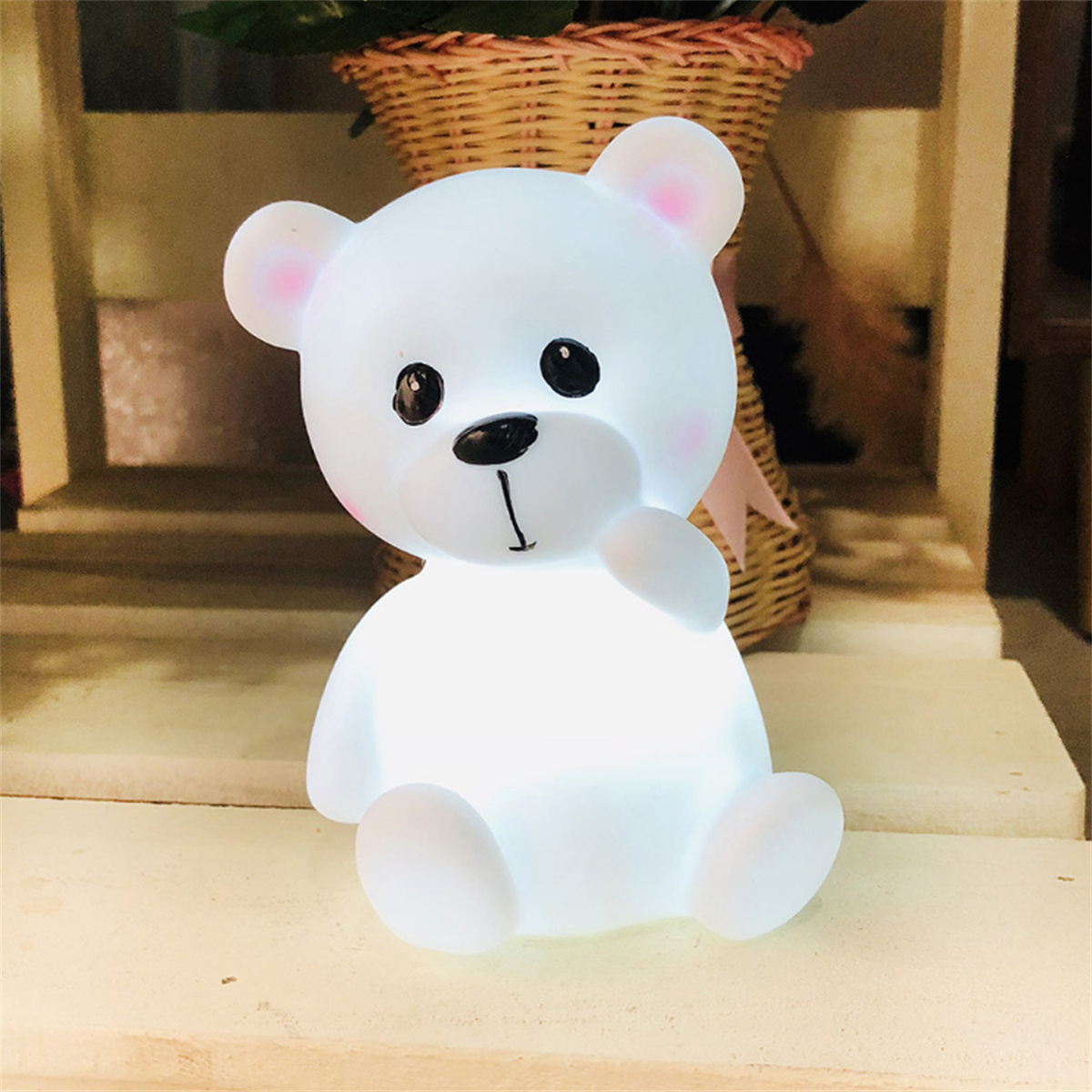 Eine leuchtende weiße Teddybär-Lampe mit rosa Wangen, schwarzen Augen und einer schwarzen Nase steht auf einer hellen Oberfläche. Im Hintergrund ist ein Weidenkorb zu sehen.
