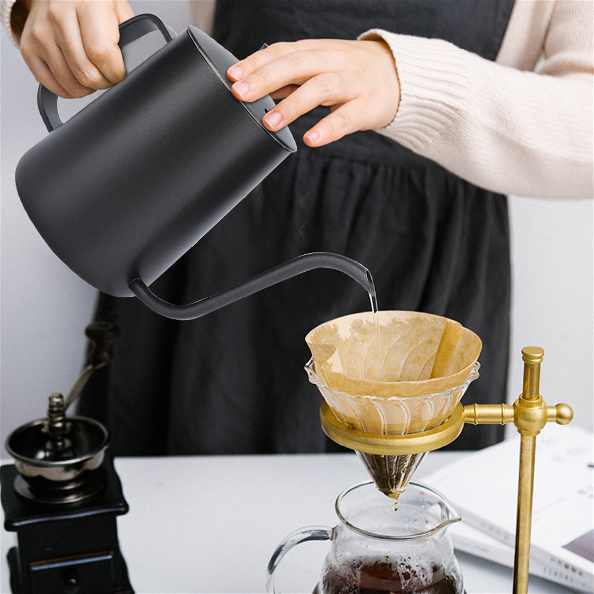 Eine Person gießt Wasser aus einem schwarzen Kessel in einen Kaffeefilter. Links befindet sich eine Kaffeemühle. Unter dem Filter eine Glaskaraffe.