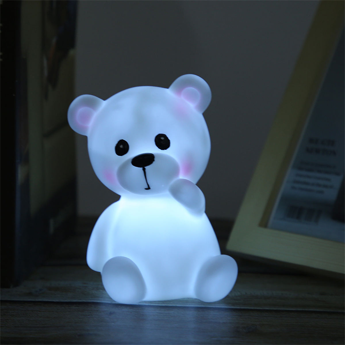 Eine leuchtende, weiße Teddybär-Lampe mit rosa Wangen und Ohren sitzt auf einer Holzoberfläche. Im Hintergrund ist ein Bilderrahmen.