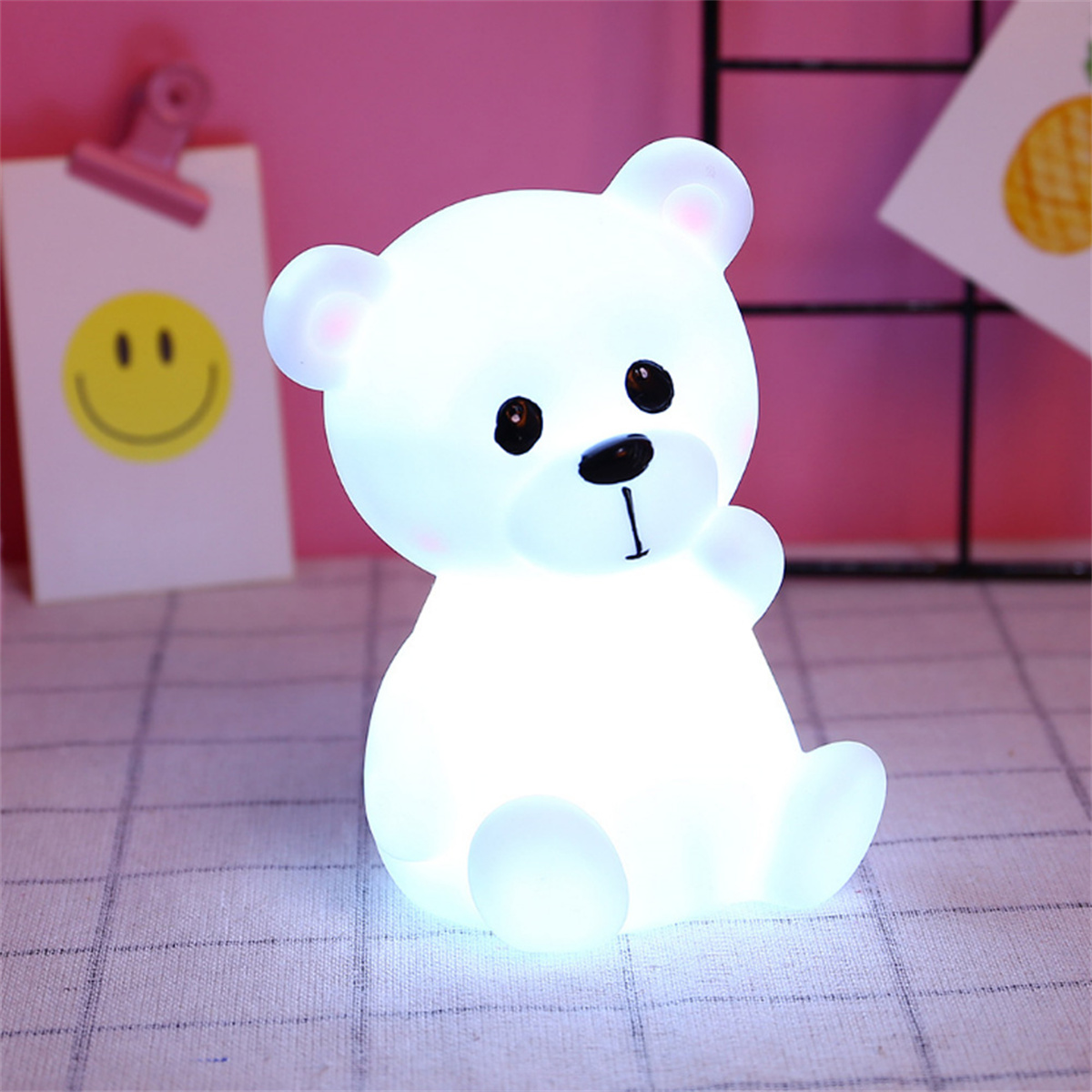 Eine leuchtende weiße Bärenlampe sitzt auf einer karierten Oberfläche. Ein Papier mit einem Smiley und rosa Hintergrund.