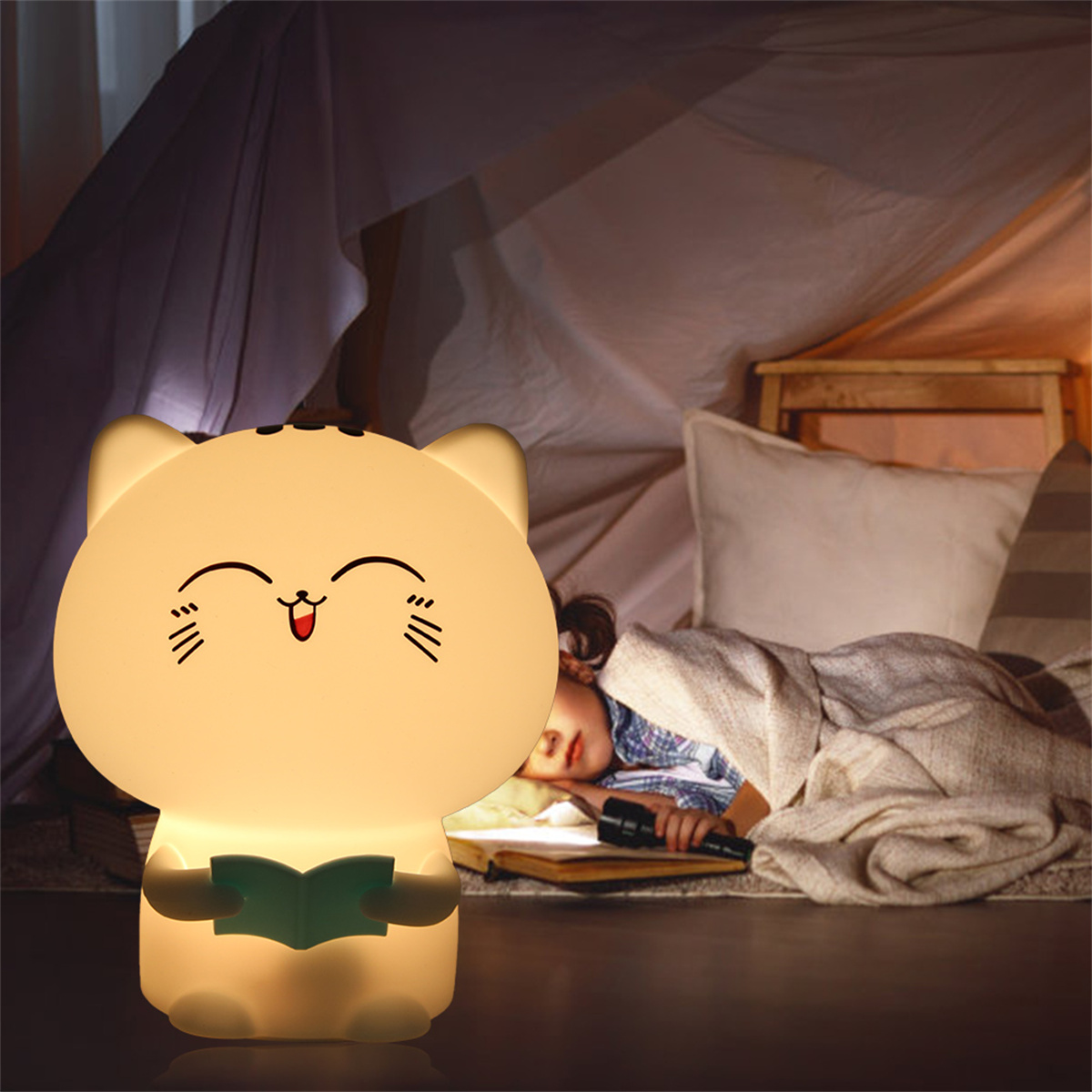 Eine katzenförmige Lampe leuchtet. Ein Kind schläft unter einer Decke in einem Zelt, neben einem Buch und einer Taschenlampe.