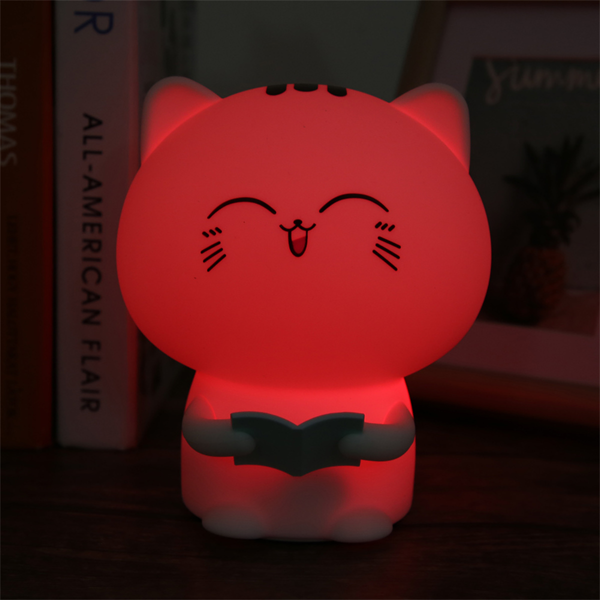 Eine leuchtende katzenförmige Lampe mit einem Buch. Sie strahlt rotes Licht aus, hat ein lächelndes Gesicht und steht auf dunkler Oberfläche.