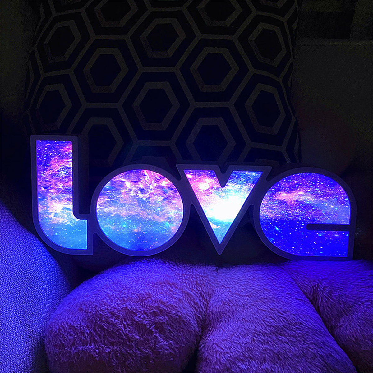 Beleuchtetes 'LOVE'-Schild mit Galaxiedesign, das auf einem pinkfarbenen Kissen liegt. Der Hintergrund ist ein gemustertes Kissen.