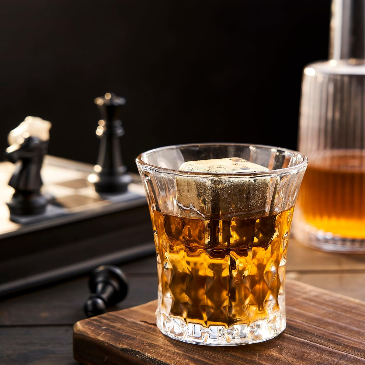 Ein Glas bernsteinfarbener Flüssigkeit mit einem Eiswürfel steht auf einem Holzbrett. Schachfiguren und eine Karaffe sind im Hintergrund.