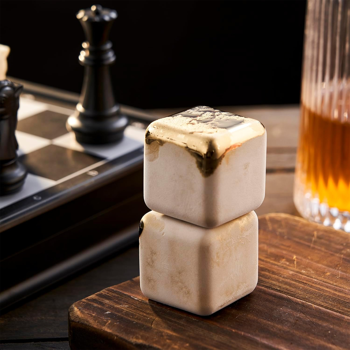 Zwei gestapelte quadratische Eiswürfel mit goldenem Rand, Schachfiguren, ein Glas bernsteinfarbener Flüssigkeit und ein Holzbrett sind sichtbar.