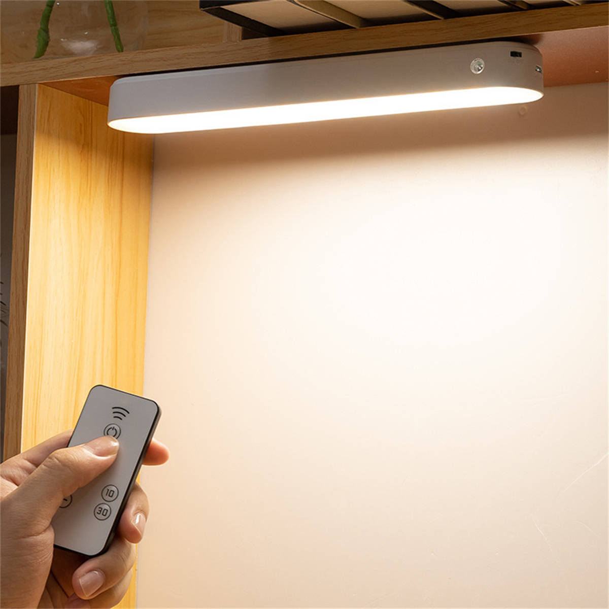 Eine Hand hält eine Fernbedienung, die auf eine weiße LED-Leuchte gerichtet ist. Das Licht ist unter einem Holzregal montiert.