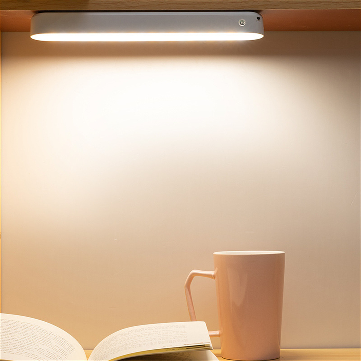 Ein warmtoniges Bild zeigt eine beleuchtete Lampe, einen rosa Becher und ein offenes Buch auf einer Holzoberfläche. Der Hintergrund ist eine Wand.