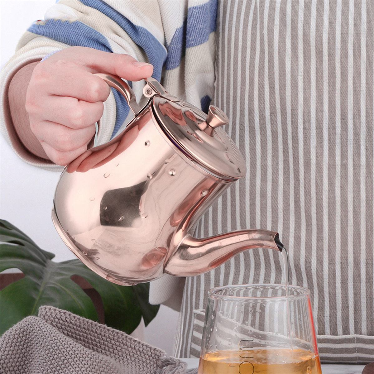 Eine Person gießt Flüssigkeit aus einer Kupferkanne in ein Glas. Die Person trägt eine gestreifte Schürze und einen Pullover. Wassertropfen auf der Kanne.
