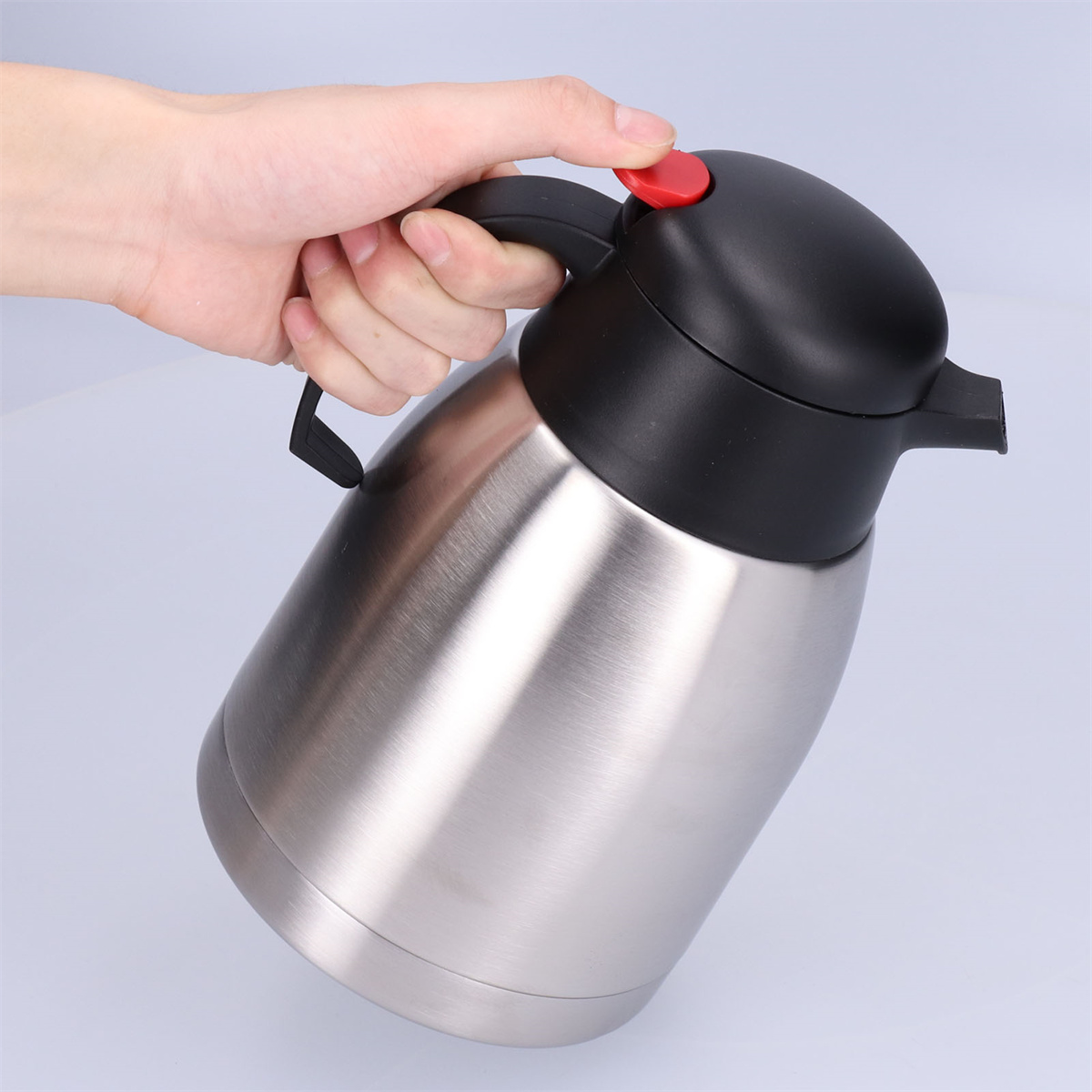 Eine Hand hält eine Edelstahl-Thermoskanne. Der Deckel ist schwarz mit einem roten Knopf. Der Hintergrund ist hellgrau.