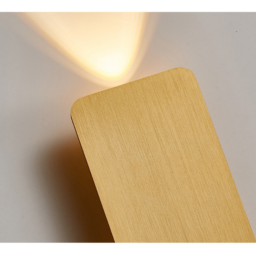 Eine goldene rechteckige Wandleuchte mit strukturierter Oberfläche. Licht scheint von oben. Der Hintergrund ist eine hellgraue Wand.