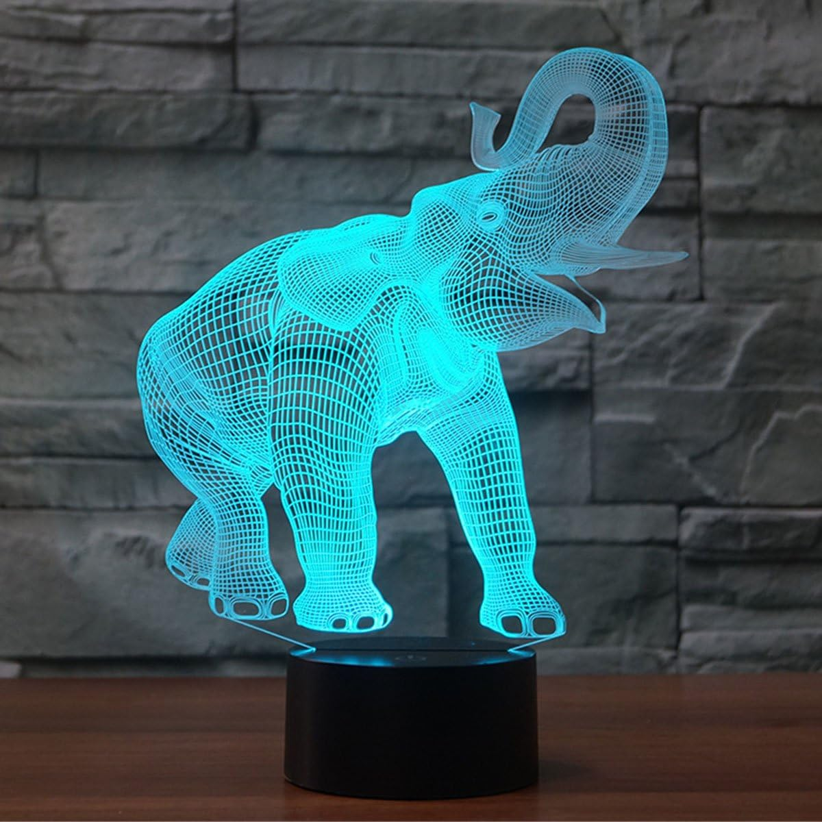 Eine blaue Drahtgitter-Elefantenlampe steht auf einem schwarzen Sockel. Der Elefant ist beleuchtet, mit Backsteinmauer im Hintergrund.