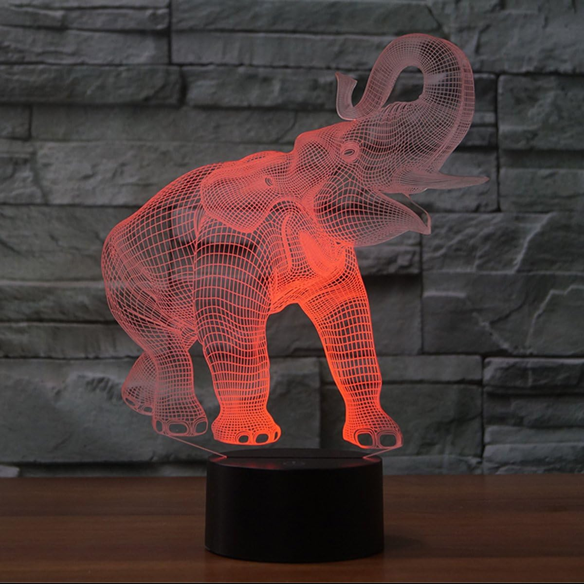 Eine rot beleuchtete Elefantenlampe steht auf einem schwarzen Sockel vor einer grauen Steinmauer. Der Elefant besteht aus einem Drahtgitter.