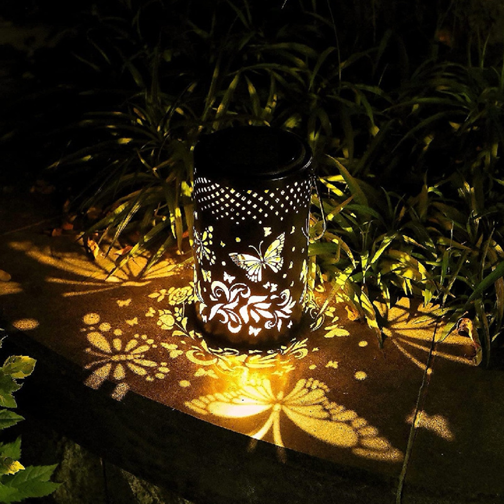 Eine schwarze Laterne mit Schmetterlings- und Blumenausschnitten wirft Licht und Schatten auf eine Steinoberfläche. Grünes Laub im Hintergrund.