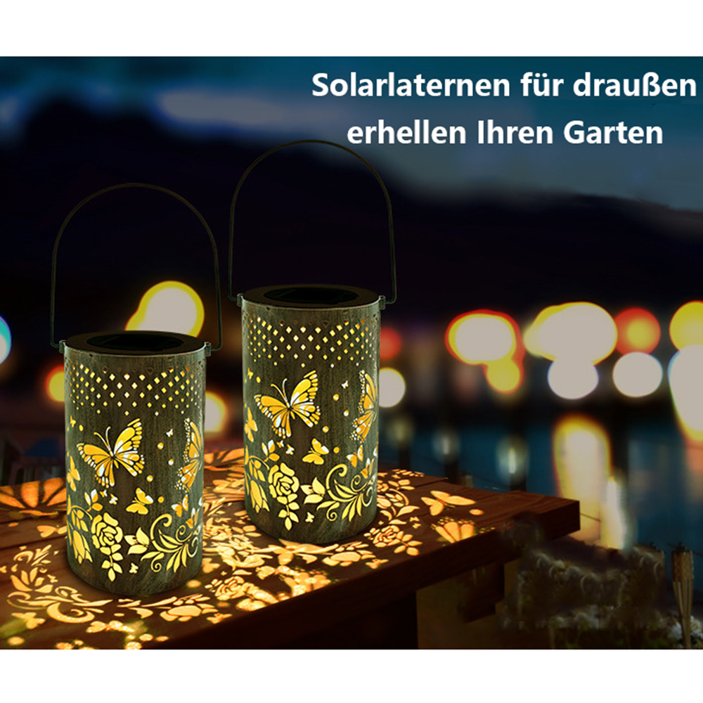 Zwei Solar-Laternen mit Schmetterlings- und Blumenausschnitten auf Holz. Nachts leuchtend. Dunkler Hintergrund mit Lichtern.