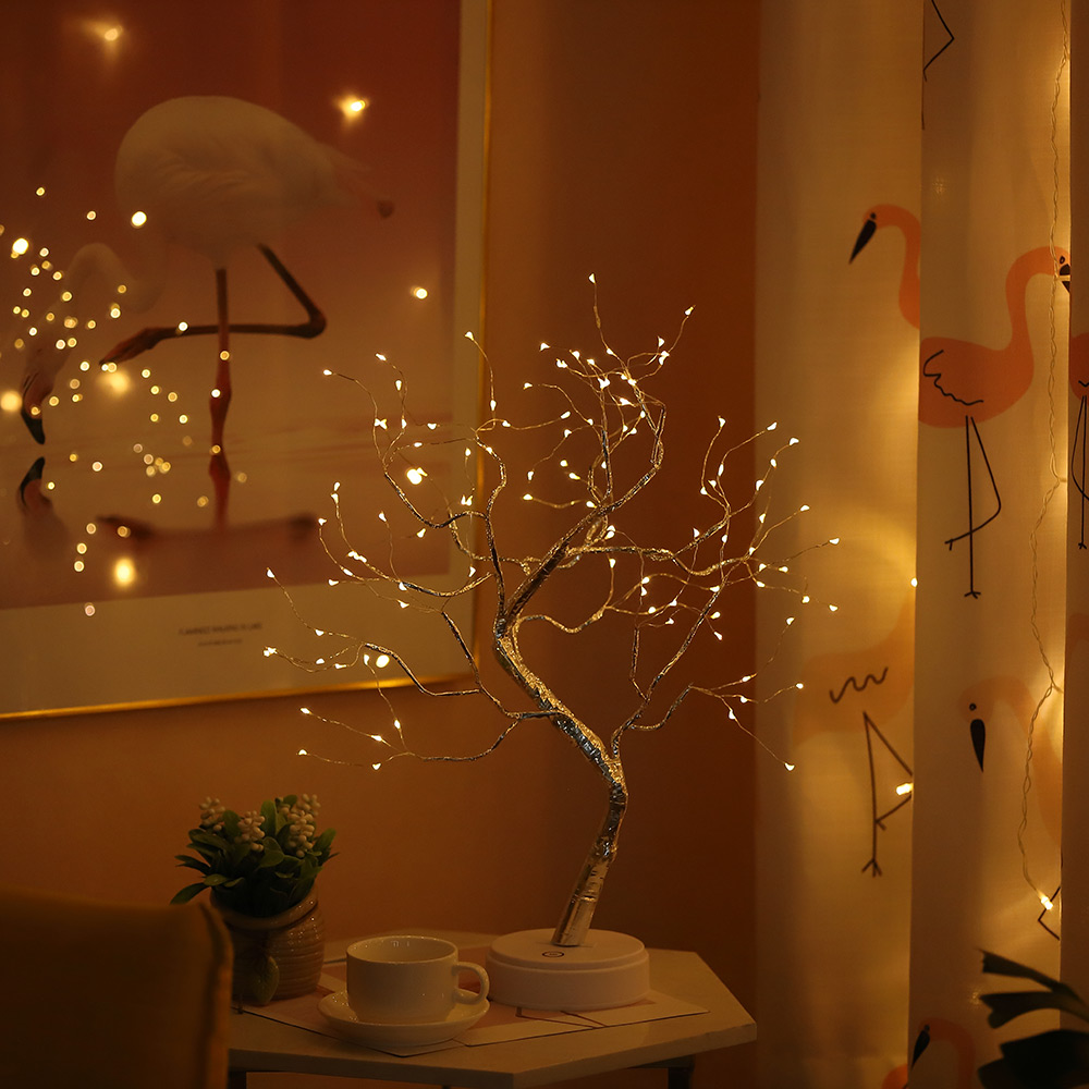 Eine dekorative baumförmige Lampe mit kleinen Lichtern auf einem Tisch. Dahinter ein Flamingodruckvorhang und ein Gemälde eines Flamingos.