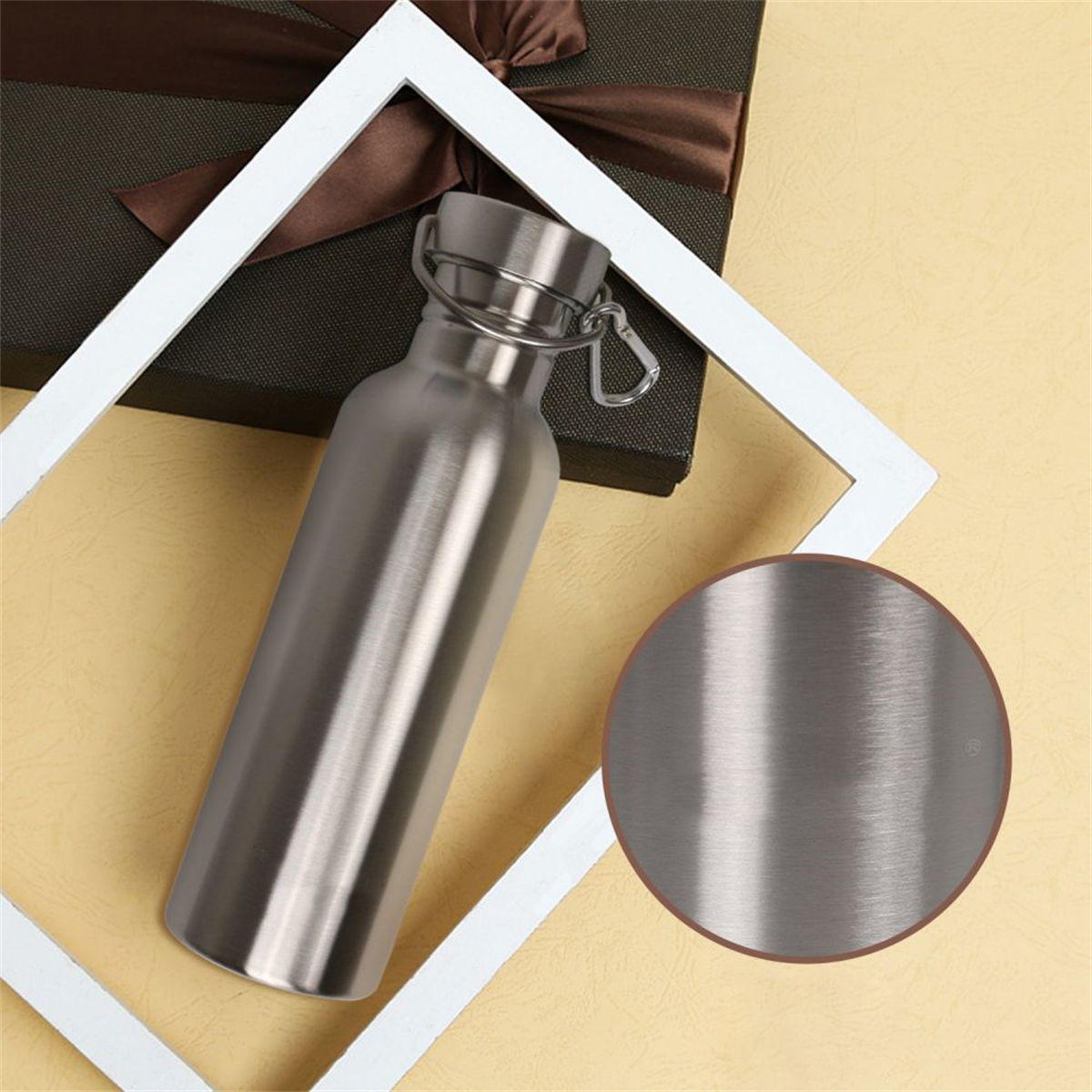 Eine Edelstahl-Wasserflasche mit silbernem Verschluss und Clip liegt auf gelbem Untergrund, neben einer Geschenkbox und einem weißen Rahmen.