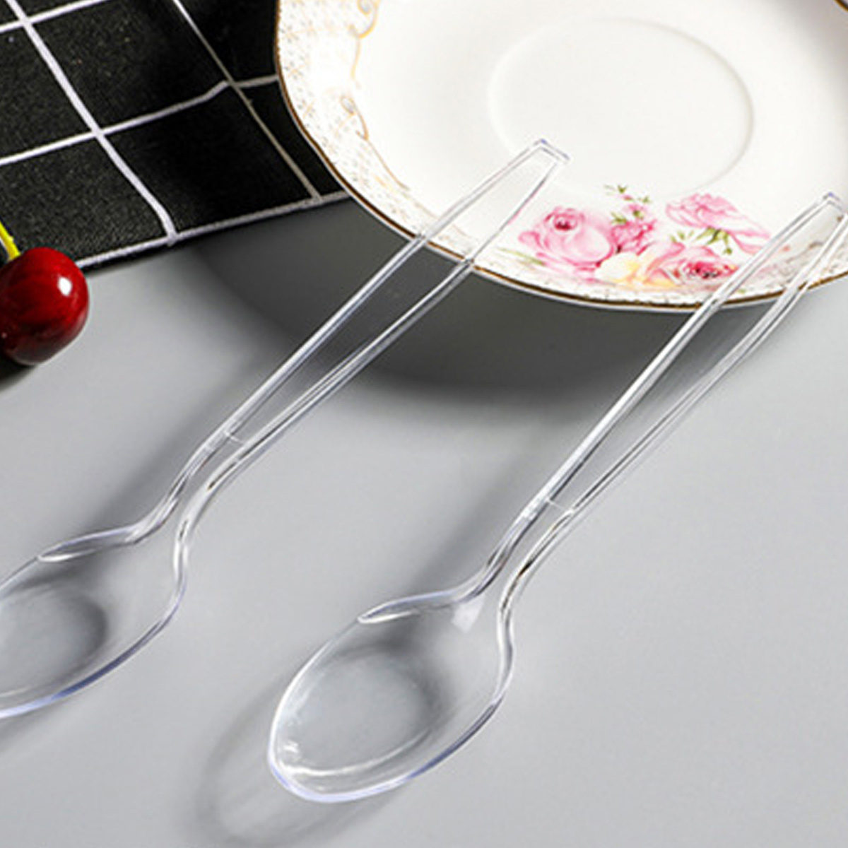 Zwei durchsichtige Plastiklöffel liegen auf einer grauen Oberfläche. Eine Kirsche und eine Blumenvase sind im Hintergrund.