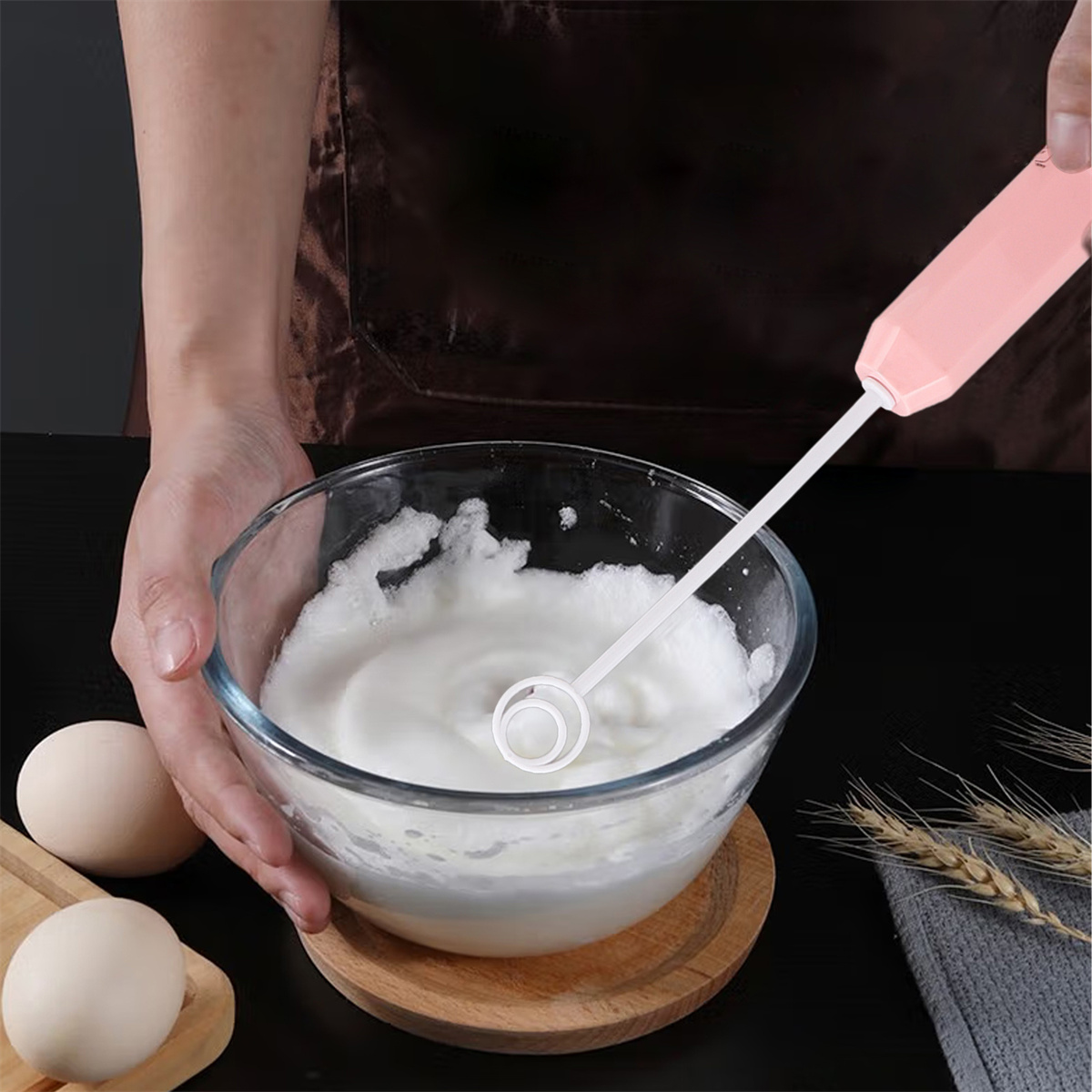 Eine Person verrührt Eier mit einem rosa elektrischen Mixer in einer Glasschüssel. Zwei Eier und Weizen liegen auf dem Tisch.