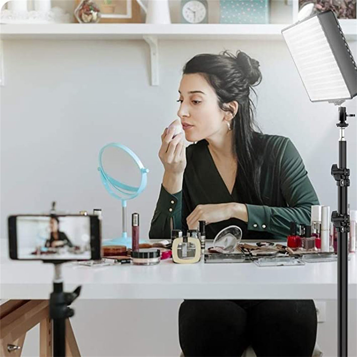 Eine Frau trägt Make-up vor einem Spiegel und Smartphone auf. Kosmetika liegen auf dem Tisch. Im Hintergrund sind Ringlicht und Regal.