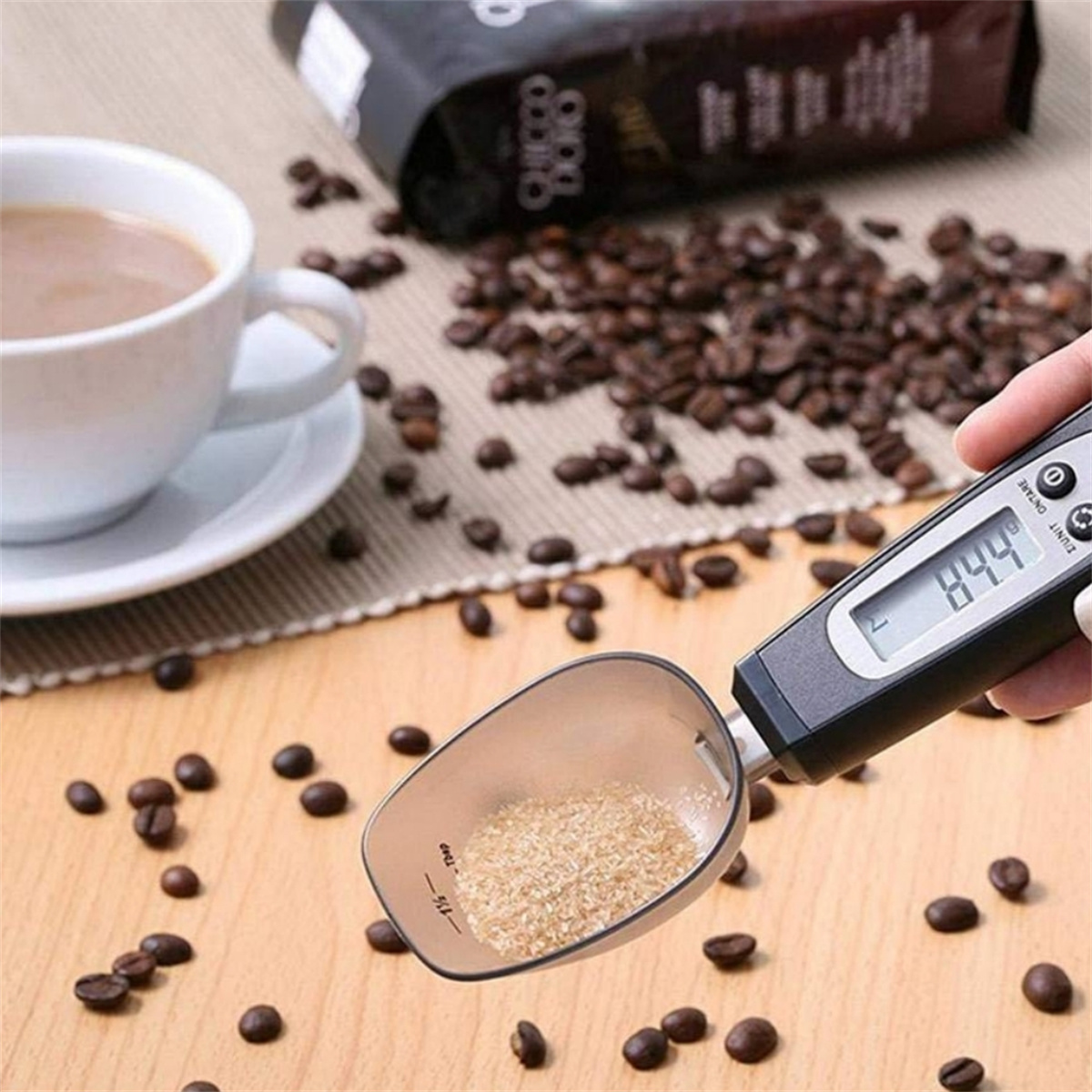 Eine Kaffeetasse und eine Löffelwaage auf einem Holztisch, umgeben von Kaffeebohnen. Die Waage zeigt 844 an, und der Löffel enthält braunen Zucker.