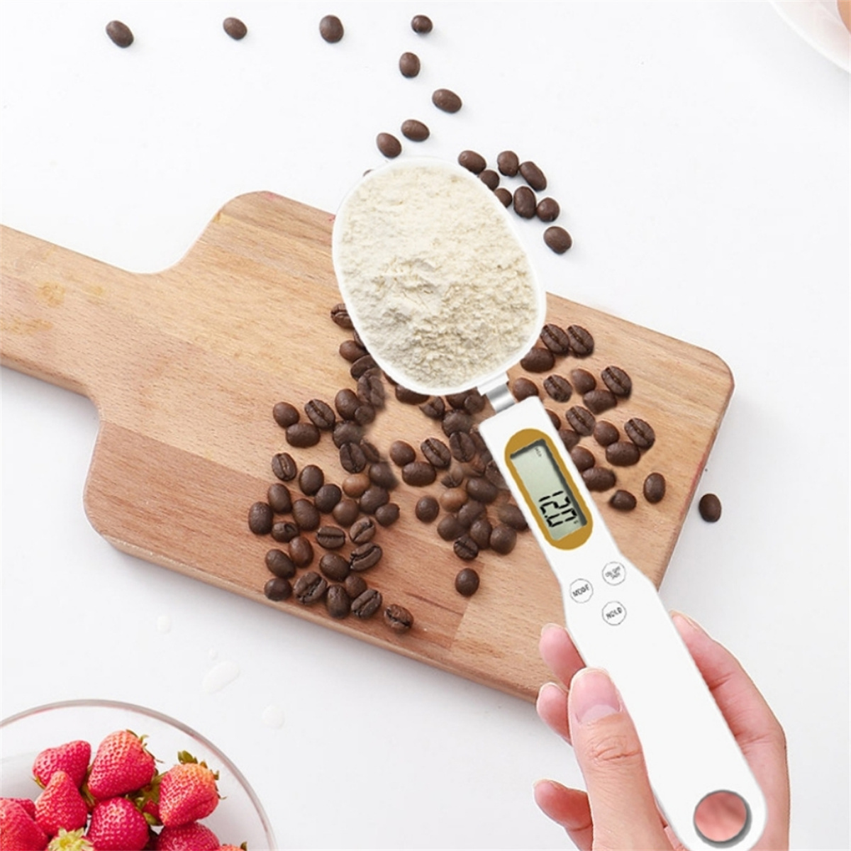 Ein weißer digitaler Küchenwaagenlöffel mit Mehl, Kaffeebohnen auf einem Holzbrett und Erdbeeren in einer Schüssel.