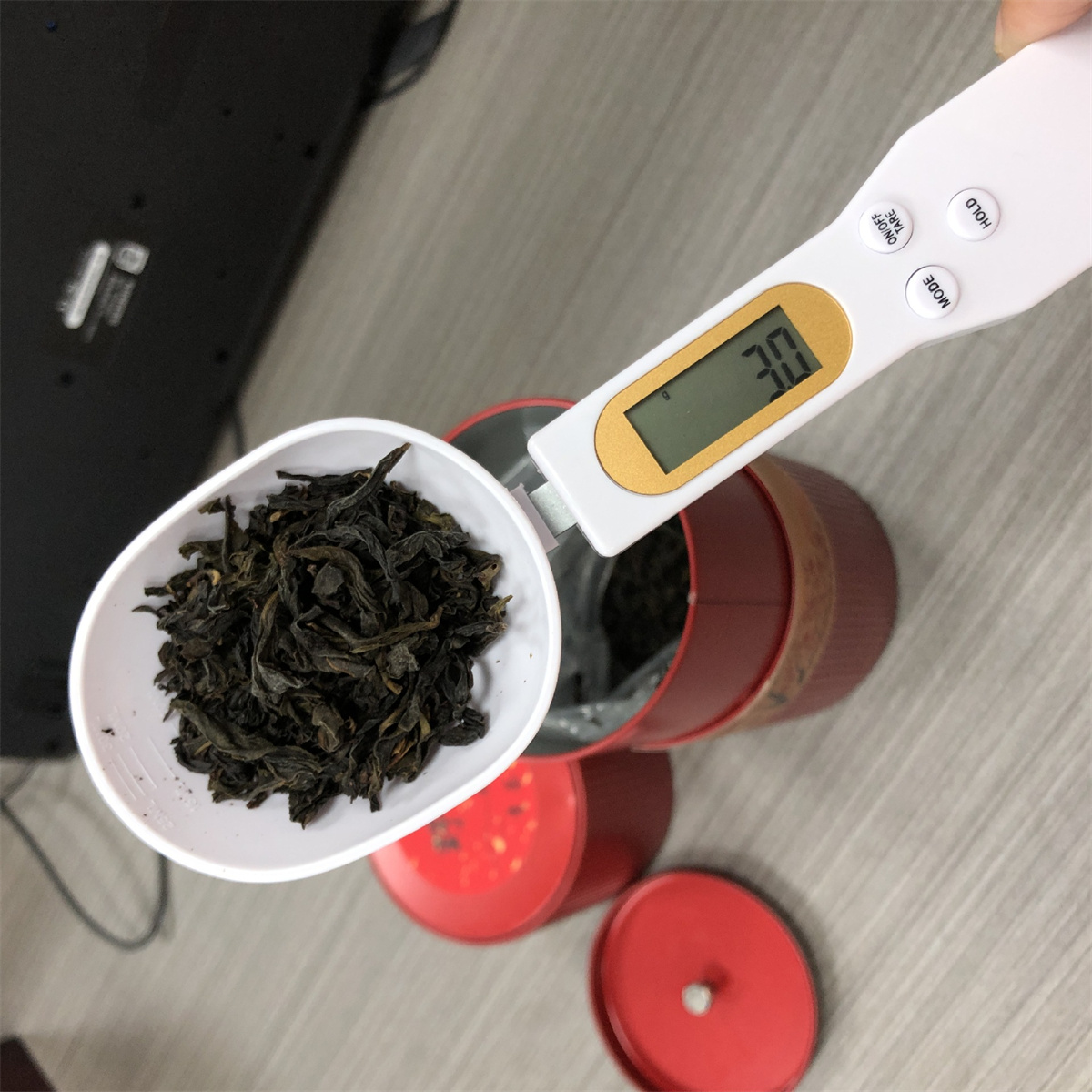 Ein weißer Löffel mit Teeblättern wird über einer roten Dose gehalten. Eine Digitalwaage zeigt 3,0 an. Ein roter Deckel befindet sich in der Nähe.