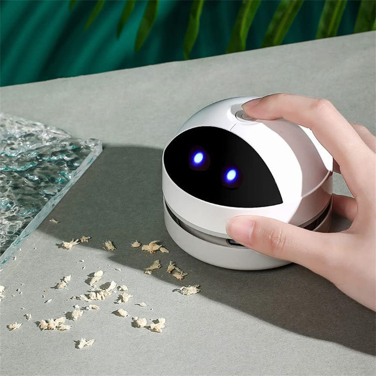 Eine Hand drückt einen Knopf auf einem weißen, roboterförmigen Mini-Staubsauger. Verstreute Trümmer und ein Glasteil sind sichtbar. Blaue Lichter leuchten.