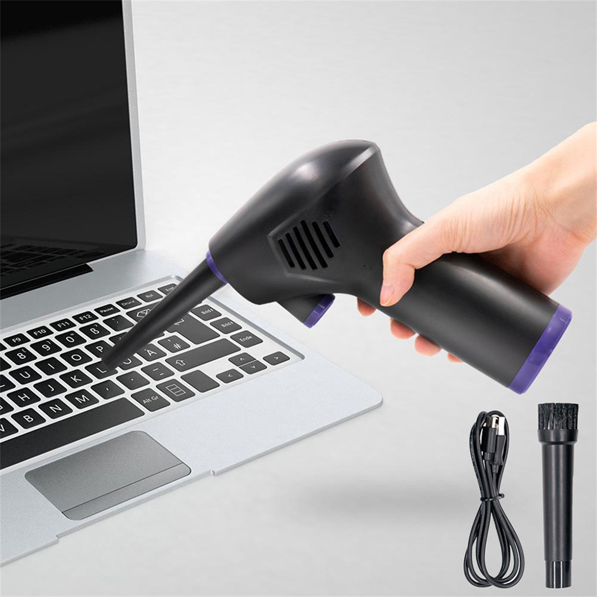 Eine Hand hält einen schwarzen elektrischen Staubwedel und reinigt eine Laptop-Tastatur. Zubehör wie Bürste und Ladekabel sind vorhanden.