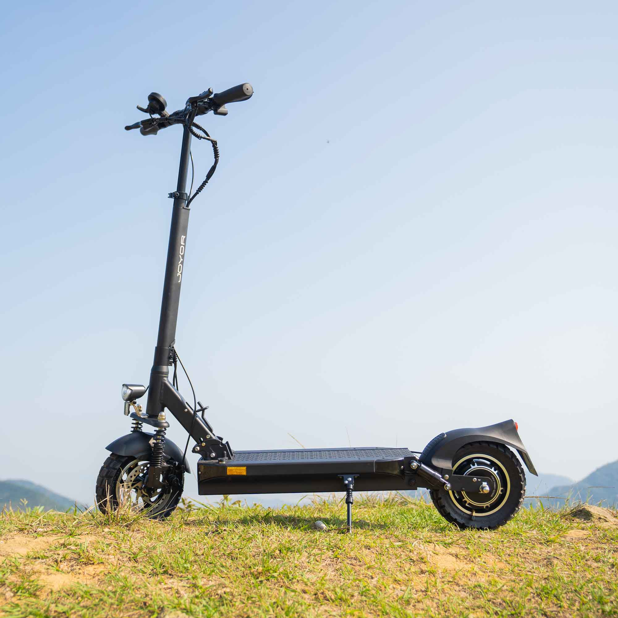 Patinete eléctrico negro sobre hierba, fondo de cielo azul. El patinete tiene faro, timbre y pie de apoyo.
