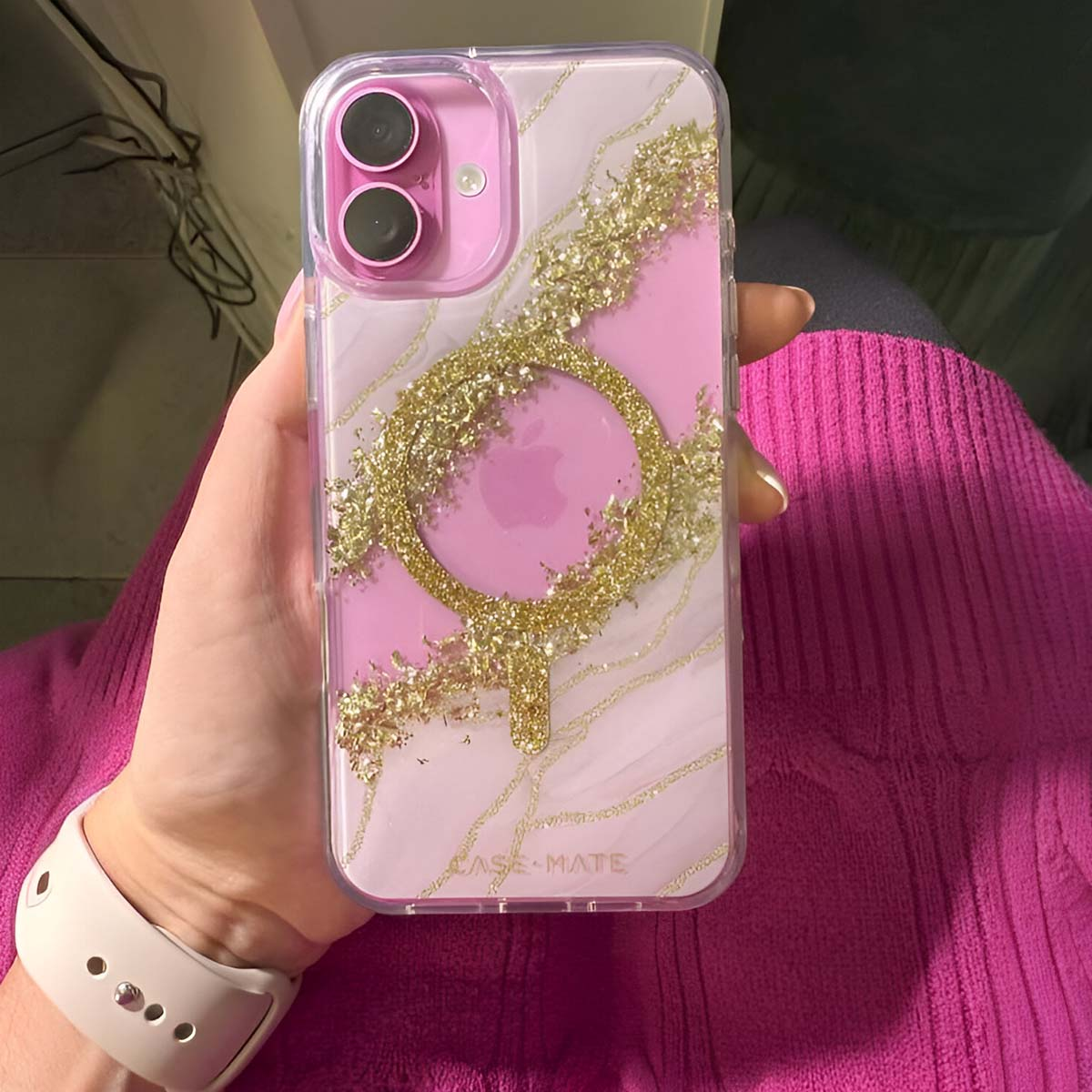 Una mano che tiene una custodia per iPhone. La custodia è rosa con glitter dorati e un bordo trasparente. L'Apple Watch è in basso a sinistra.