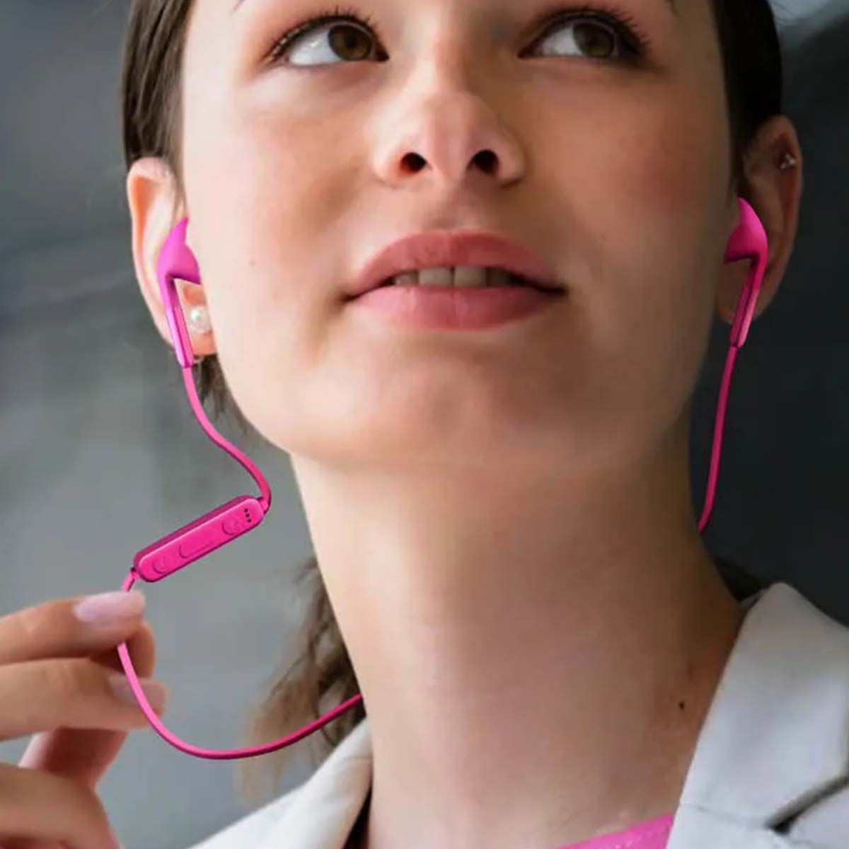 Una donna con auricolari rosa, che guarda in alto. Indossa un blazer bianco. Gli auricolari hanno un modulo di controllo.