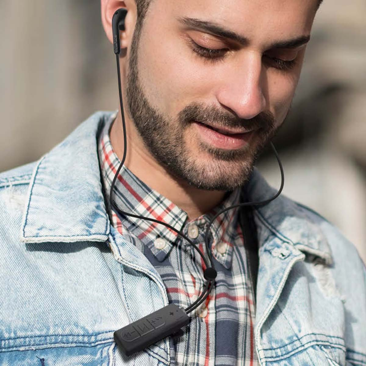 Un uomo con una giacca di jeans e una camicia a quadri utilizza auricolari wireless neri. Ha la barba e gli occhi chiusi.