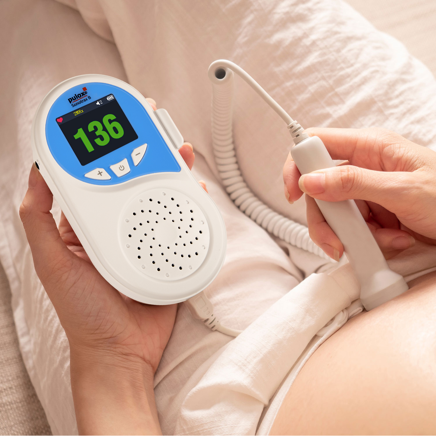 Eine Person hält ein fetales Dopplergerät auf einem schwangeren Bauch. Das Gerät zeigt eine Herzfrequenz von 136 an. Der Hintergrund ist ein Bett.