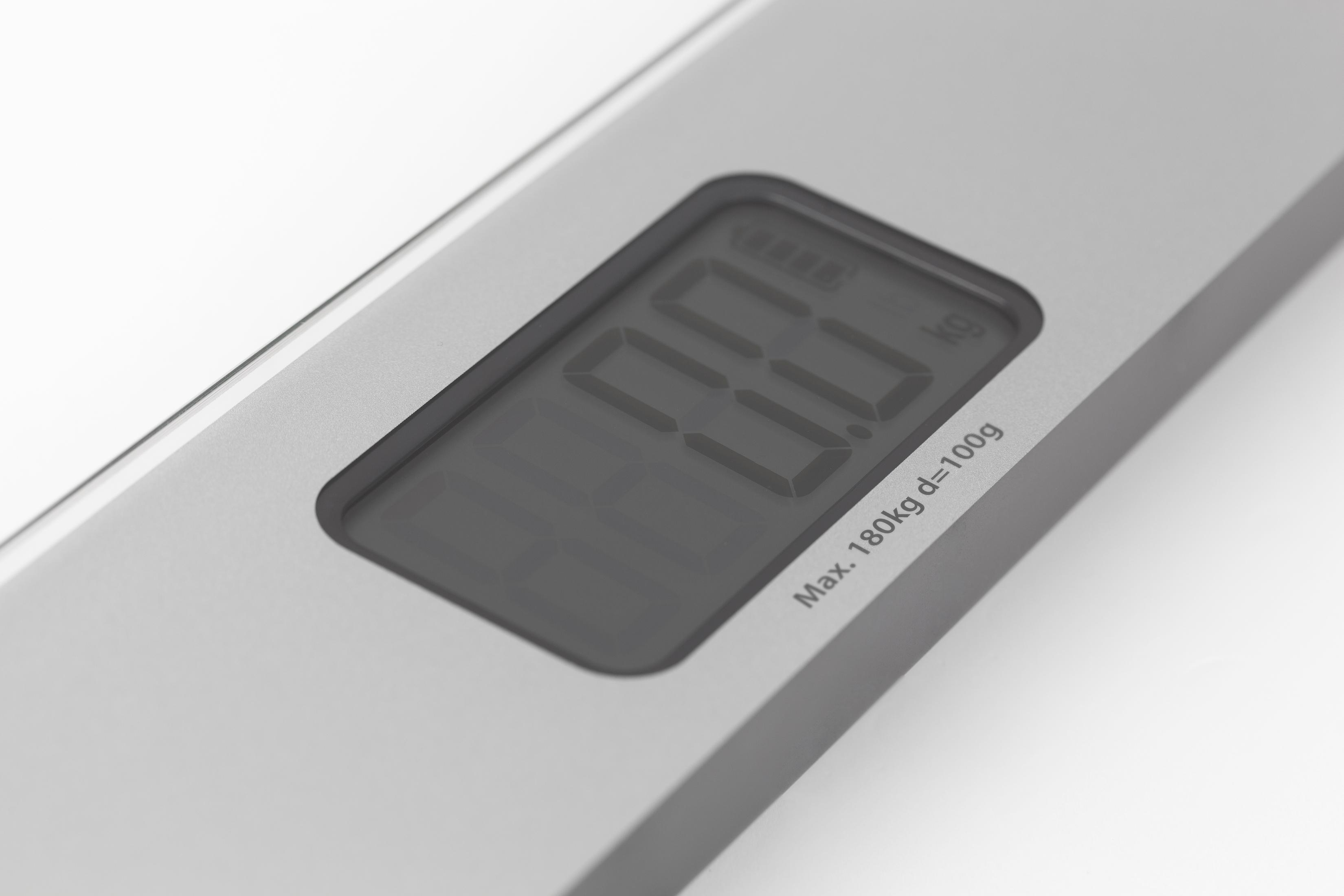 Eine Digitalwaage zeigt 0,0 kg an. Die Waage ist silberfarben, der Hintergrund weiß mit Text: Max. 180 kg d=100 g.