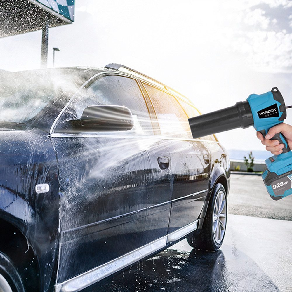 Eine Person trocknet mit einem blau-schwarzen Gebläse ein schwarzes Auto. Wasser spritzt vom Auto in der Sonne.