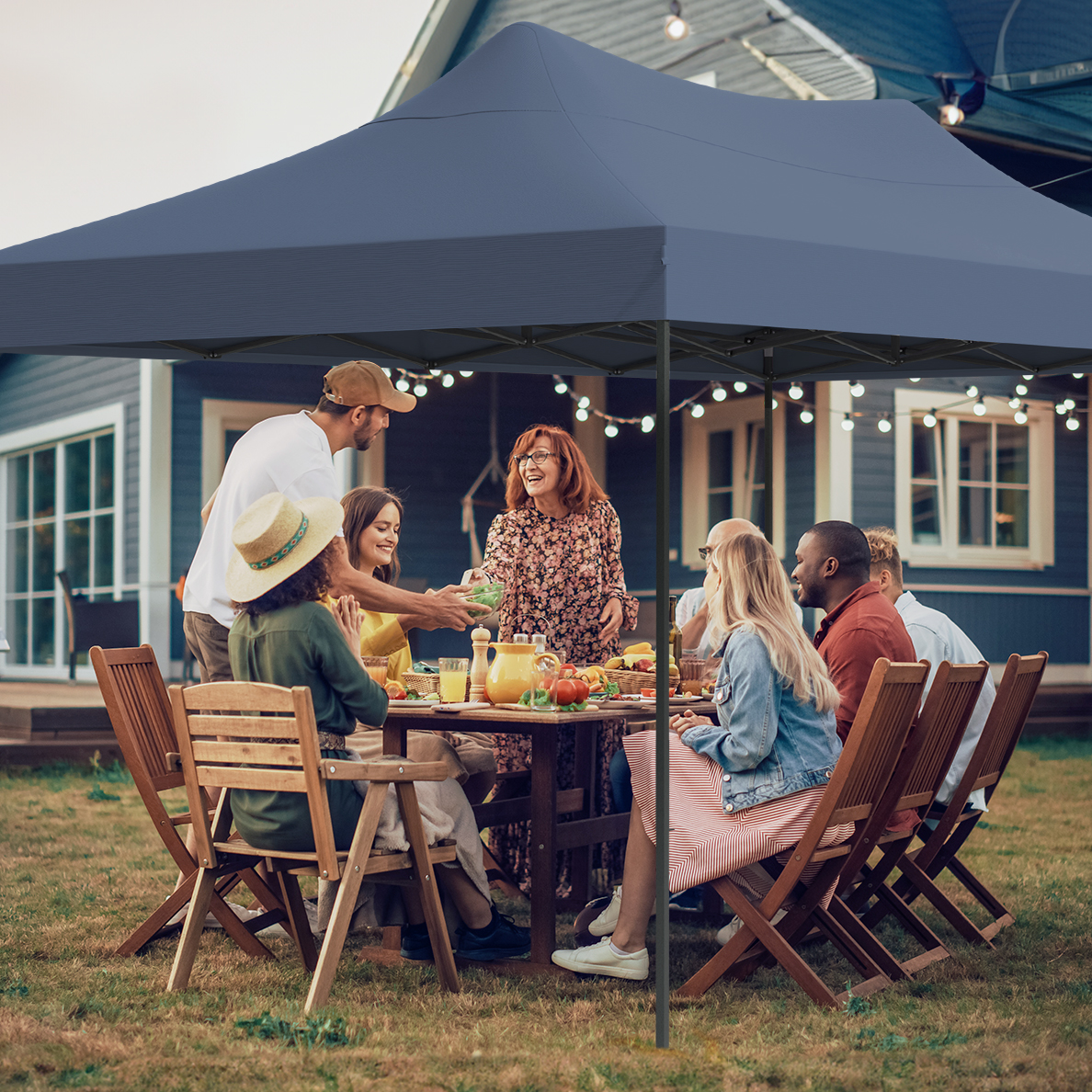 Group of people at a backyard dinner party under a blue canopy. They are seated around a wooden table with food and drinks.