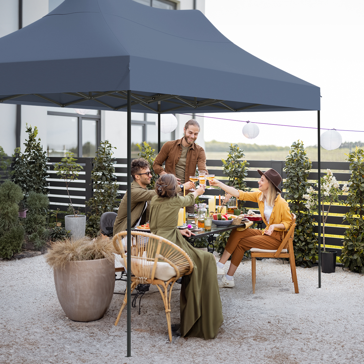 Group of friends toasting at an outdoor table, under a canopy. Food and drinks are on the table. Background includes a house and plants.